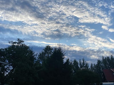 The image depicts a view of the sky above trees and a rooftop in Leipyasuo, Russia. The sky is a mix of blue and white, with scattered clouds. The clouds appear to be thin and wispy, with some areas of sunlight breaking through. The trees in the foreground are dark green and silhouetted against the sky. A portion of a red-tiled roof with a ladder is visible on the right side of the image. There are no people or animals present in the scene. The overall impression is one of a natural landscape under a cloudy sky.