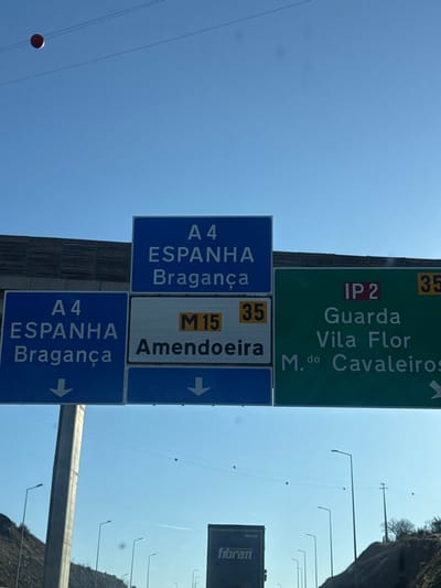 The image displays an overhead highway sign gantry on a clear, bright blue day, indicating directions at a road junction near Macedo de Cavaleiros, Portugal.
The gantry supports multiple directional signs. From left to right, above the lanes:
1. A blue sign reads "A 4 ESPANHA Bragança."
2. Below it, another blue sign reads "A 4 ESPANHA Bragança" with a downward arrow, indicating an exit.
3. Adjacent to the lower A4 sign, a white sign states "M 15 35 Amendoeira" with a downward arrow, indicating an exit for Amendoeira via M15, 35 units away.
4. To the right, a green sign reads "IP 2 35 Guarda Vila Flor M.do Cavaleiros" with a rightward arrow, indicating an exit for IP2 towards Guarda, Vila Flor, and Macedo de Cavaleiros, 35 units away.
The perspective is from a vehicle on the highway. In the lower background, a dark blue commercial truck displaying "fibran" on its trailer is partially visible. Multiple dark streetlights line the roadway, and sparse vegetation covers the adjacent hillsides. Overhead, several power lines traverse the sky, featuring red spherical markers (aircraft warning spheres), with one prominent sphere in the upper left. The scene is an interchange where the IP2 provides an exit option to Macedo de Cavaleiros.