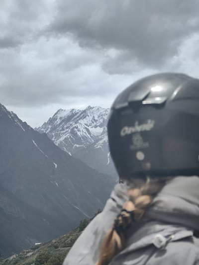 The image shows a close-up, over-the-shoulder view of a person wearing a black helmet and a grey jacket. The person's braided blonde hair is visible. The helmet has a white logo and some writing on it. In the background, a vast mountain range is visible, with snow-capped peaks under a cloudy sky. The mountains appear to be in a remote, possibly mountainous region. The scene is likely in India, given the possible Indian certification mark on the helmet.