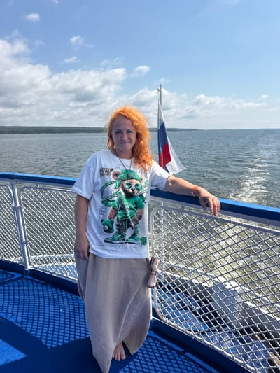 A woman with vibrant red hair stands on the deck of a boat, looking towards the camera. She is wearing a white t-shirt with a graphic print of a bear in a green tracksuit and sneakers, and a long, beige skirt. The woman is leaning against the blue railing of the boat, and her right arm, adorned with an orange wristband, is resting on it. Behind her, a Russian flag flutters on a flagpole. The boat is on a body of water, likely a river or lake, with a forested shoreline visible in the distance. The sky is blue with scattered clouds, and the water is rippled, indicating movement. The deck of the boat has a blue surface with a patterned matting. The overall impression is a pleasant day trip on the water. The location is not specified as the city of Oryol, Russia.