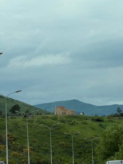 The image depicts a landscape with a cloudy sky dominating the left side of the frame. A mountain is visible in the background, partially obscured by the sky. On the right side of the image, there is a grassy hillside. A building with a red roof sits atop the hillside. A row of streetlights extends along the bottom right of the image. The location is likely Mrgvali Chala, Georgia.
