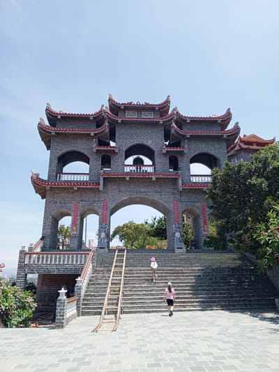 The image depicts a stone archway with multiple tiers and curved, tiled roofs, characteristic of traditional East Asian architecture. Two individuals, one wearing a pink hat and the other a pink shirt and white hat, are ascending a wide stone staircase leading to the archway. A wooden ladder is propped against the stairs to the right. Lush green trees are visible to the right and behind the structure, and pink flowers bloom on the left. The sky above is clear and blue. The scene is likely situated in Ninh Chu Ward, Vietnam, known for its historical sites and natural beauty.