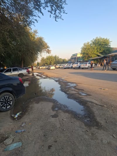 The image shows an outdoor scene in the city of Ko`kdala, Uzbekistan, featuring a street with parked cars and a large puddle of murky water. Several cars are parked along the side of the asphalt road, including a silver sedan and a black car in the foreground. A person in a brown shirt stands near the puddle, and a group of three men are standing together further down the street. Trees line the left side of the road, and in the background, there are buildings with awnings and more trees. The ground is uneven and covered with gravel and debris, including several plastic bottles scattered around the puddle. The sky is clear and blue, indicating daylight.