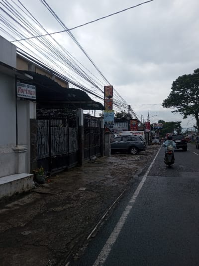 The image depicts an urban street scene under an overcast sky. A dark asphalt road, marked with a solid white line on its left, extends from the foreground into the distance. A single individual, wearing a helmet and a light-colored backpack, rides a grey scooter away from the viewer on the road.
To the left of the road, a dark, uneven, and somewhat damp sidewalk or unpaved area borders a row of structures. Immediately to the left, a white building segment displays a sign stating "LAUNDRY & DRY CLEAN FORTUNA" and "Pusat Cuci Sepatu, Karpet, dll," indicative of a laundry and specialty cleaning service. Adjacent to this building is a dark metal gate. Further along the street on the left, a dark grey SUV is parked near the curb. Beyond it, multiple commercial signs are visible, including those with the "TELKOM" logo and "YAKULT" branding, among other advertisements.
Numerous black utility wires are strung overhead, traversing the cloudy sky. On the far right, a large green tree is partially visible, providing a natural element to the urban environment. Additional vehicles are present on the road and parked further in the distance. The scene is located in Kamji Na-Yekha, Bhutan.