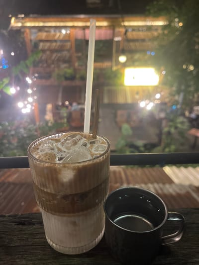 The image depicts an evening scene, likely within a cafe or garden setting, in Cần Thơ, Vietnam. In the immediate foreground, a tall, fluted glass contains a layered iced coffee drink, characterized by visible strata of white milk, brown coffee, and a foamy top layer, interspersed with ice cubes. A clear plastic straw and a slender metal spoon are present within the glass. To its right, a dark metallic cup with a handle rests on a rustic wooden surface. A dark horizontal railing crosses the frame above the wooden surface. The blurred background reveals an outdoor environment illuminated by string lights on the left and a prominent glowing rectangular yellow sign to the right. Structural elements include a multi-level wooden building adorned with plants, and corrugated metal roofing is visible beneath the railing. No individuals or specific human actions are discernible within the frame; the scene is focused on the beverages and ambient environment.