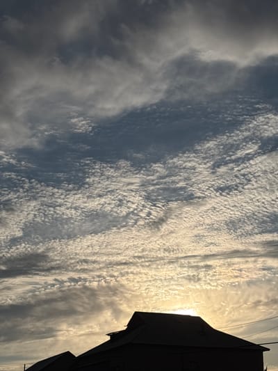 The image captures a silhouette of a building's roof against a sky filled with clouds at what appears to be sunset. The sky displays a mix of dark and light clouds, with a bright area near the horizon suggesting the sun is low. The building's dark roofline is sharply defined, with visible architectural details like gables and possibly vents. The scene appears to be set in the rural area of Жібек жолы ауылдық округі, Kazakhstan, given the context provided, and the architecture is consistent with a dwelling or similar structure in such a location. There are no people or discernible actions depicted in the image.
