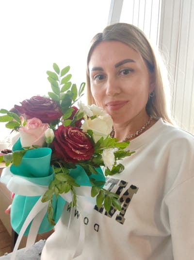 A woman with blonde hair and fair skin holds a bouquet of roses. She is wearing a white sweatshirt with a leopard print graphic and a silver necklace. The bouquet is decorated with teal and white ribbons. The background appears to be indoors with vertical blinds. The image is a selfie taken in Amursk, Russia.