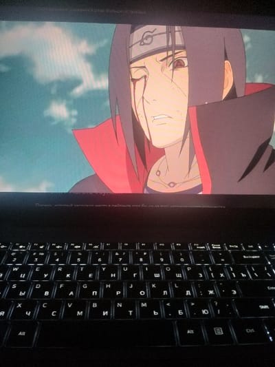 The image displays a laptop, angled to show both the screen and the keyboard. On the screen is a close-up of the anime character Itachi Uchiha. The keyboard is dark, with white illuminated letters in Russian. The keyboard labels include 'Alt', 'Ctrl', 'Enter', the Russian alphabet, and various symbols. The scene is dimly lit, emphasizing the glowing keyboard and the illuminated laptop screen.