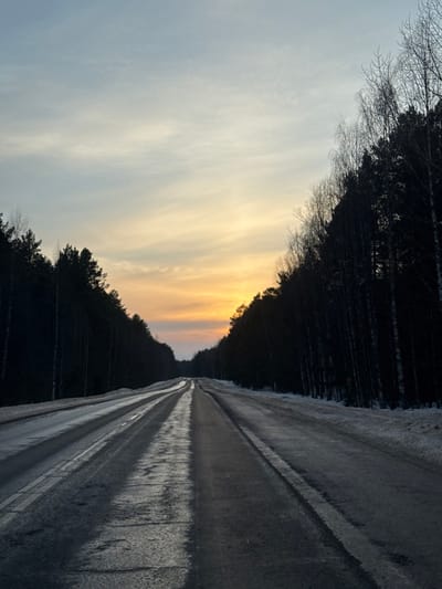 An image depicts a two-lane asphalt road traversing a dense forest during what appears to be either a sunrise or sunset. The sky displays a gradient from light blue at the zenith to warm orange and yellow hues near the horizon, with subtle cloud formations. The road surface is partially wet and reflective, suggesting ice or moisture, with visible tire tracks in the center and along the lanes. Faint white lane markings are discernible. Snow covers the shoulders of the road. On both sides, tall, dark silhouettes of evergreen and deciduous trees (many appearing bare) form a continuous, dense barrier, extending into the distance. The road itself is relatively straight but shows some undulations as it recedes from view. No individuals, vehicles, or specific man-made structures other than the road are visible. The environment indicates a winter setting. This scene is observed within the broader vicinity of Зюкайка, Russia.