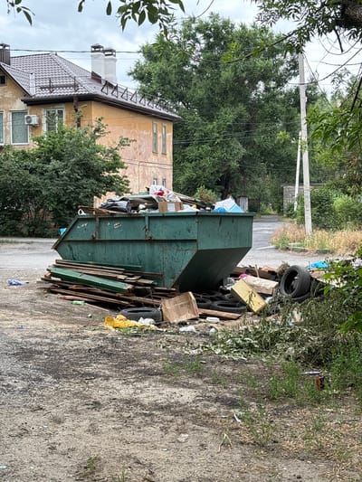 The image depicts an exterior scene, likely in Novocherkassk, Russia, characterized by a large, overflowing green dumpster filled with various types of refuse. The dumpster is situated on a gravelly, unpaved surface, indicative of an industrial or service area, or a less maintained residential zone. Surrounding the dumpster, a significant amount of debris has accumulated, including wooden planks, cardboard boxes, discarded tires, and plastic bags. The scene also includes a building with a light-colored facade and a tiled roof, partially obscured by trees. The overall impression is one of neglect and accumulation of waste.