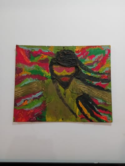A painting is mounted on a white wall. The painting features thick, textured strokes of color. Dominant colors include red, green, yellow, brown, and black. The overall impression is abstract and somewhat crude, with no identifiable subject matter clearly defined. The location is Chopdem, India.
