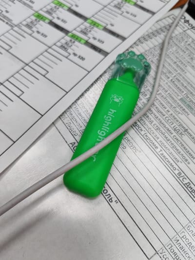 A green vape pen with the text "highligh" is placed diagonally across a document with columns of text. The vape pen has a transparent cap shaped like a paw print. A white cable is coiled around the vape pen and extends towards the bottom left of the image. The document appears to be a printed form or report with lines and printed text in Russian. The background is a light-colored, textured surface, possibly a desk. The image is taken from a slightly elevated angle, looking down at the objects. The lighting suggests an indoor environment. The text on the document includes what appears to be company names, locations, and numerical data. The text "Иланск" and "Бородино" are visible on the document, suggesting the scene is related to Borodino, Russia.