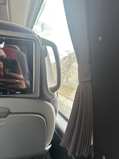 The image shows the interior of a bus, likely in Spain. The location, based on the coordinates, is a highway near La Losa, specifically the A-1 highway. The scene is captured from the perspective of a passenger seated by the window, looking out. The passenger's reflection is visible in the seatback screen. The window is partially covered by a gray curtain. The bus seat and its armrest are visible in the foreground. The surrounding landscape, seen through the window, is a rocky terrain with a highway in the distance. The environment is characterized by daylight, a cloudy sky, and a sunny atmosphere. The scene suggests a journey underway, with the passenger seated by the window.