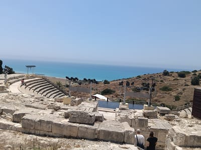 In Episkopi, Cyprus, the image displays the ruins of an ancient amphitheater overlooking the sea. The foreground is dominated by large, rough-hewn stone blocks, indicating the archaeological nature of the site. Beyond the ruins, curved stone seating tiers of the amphitheater are visible, with a single person sitting on the steps. Further back, a stage area is set up with lighting equipment and a white tent. The Mediterranean Sea forms a vibrant blue backdrop, meeting the hazy horizon under a clear sky. The surrounding landscape is dry and rocky, with scattered bushes and trees. Two individuals, viewed from behind, are walking away from the camera and towards the ruins, one wearing a white long-sleeved shirt and a cap, the other in a black t-shirt. The overall scene captures a historical site in a coastal environment on a bright, sunny day.