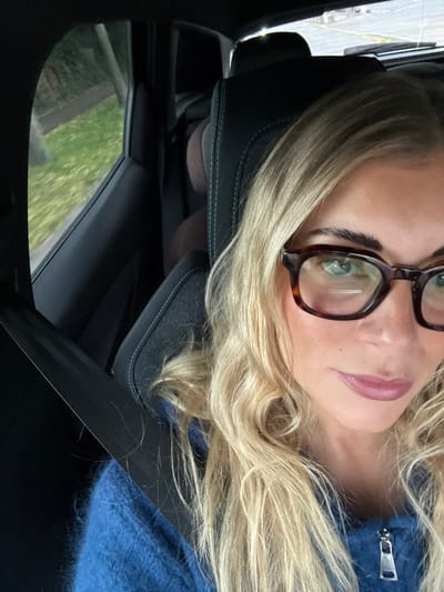 An image captured in the city of Trets, France, depicts a female seated in the front passenger seat of a vehicle. She has blonde, wavy hair and is wearing dark, rectangular, tortoise-shell framed eyeglasses, revealing light-colored eyes. Her lips are a muted pink-mauve tone. She is dressed in a blue, fuzzy, zippered garment. A black seatbelt is visible across her chest and left shoulder. The car's interior features a dark gray fabric seat with light stitching, and dark interior trim. In the background, a dark brown padded headrest of another seat is visible. Through the passenger window, green foliage and what appears to be a light-colored ground or road surface are discernible, suggesting an outdoor environment.