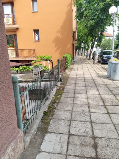 The image depicts a sidewalk in Belogradchik, Bulgaria, adjacent to an orange building. The sidewalk is paved with square concrete tiles and has a slight incline. Trees line the sidewalk on the right side, casting shadows. Several cars are parked along the street next to the sidewalk. Two large, grey trash cans are situated on the sidewalk. A green metal fence runs along the left side of the sidewalk, bordering a residential area with potted plants and a balcony visible on the orange building. The overall scene suggests a typical urban streetscape.