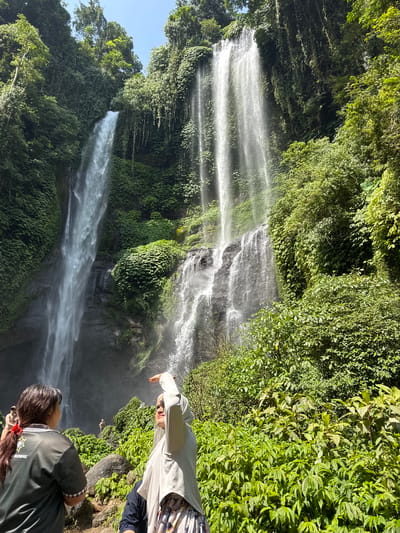 The image captures a view of the Sekumpul Waterfalls in Sawan, Indonesia. In the foreground, two women are visible. The woman on the left, wearing a dark green shirt, faces away from the camera and towards the waterfalls. She has dark hair tied back with a red ribbon. The woman on the right, wearing a light grey hijab and a patterned skirt, is facing the camera with her right arm raised, seemingly shielding her eyes or gesturing. Behind them, lush green vegetation and large rocks form the immediate landscape. In the mid-ground, more people are visible, including a person standing in the mist of the waterfall. The main subjects are the towering Sekumpul Waterfalls, which cascade down a steep, verdant cliff face. The water is bright white as it falls, creating a mist at the base. The surrounding environment is dense with tropical foliage, including trees and various types of plants, all bathed in bright sunlight. The sky is clear and blue.