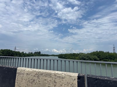 The image shows a view from a bridge over a body of water, likely a river or lake, with lush green trees lining the banks. The sky is partially cloudy with a mix of blue sky and white cumulus clouds. In the foreground, a concrete barrier with black and white paint markings separates the viewer from the railing of the bridge. The railing is made of vertical metal bars. In the background, on the left side, a tall electricity pylon can be seen amongst the trees. On the right side, another, more distant electricity pylon is visible. The water appears calm and a light turquoise color. The scene suggests a natural or semi-natural environment within or near the city of Ghvritkhevi Community, Georgia, possibly during daylight hours under a partly sunny sky. No people are visible in the image.