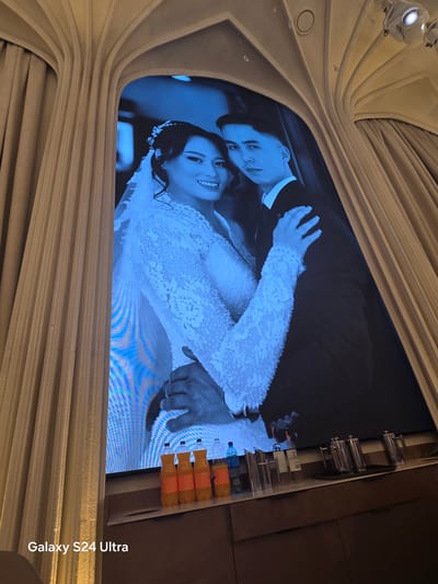 The media file is a photograph of a large screen displaying a wedding photo. The photo features a bride and groom, both dressed in formal attire. The groom is wearing a dark suit and a white shirt, while the bride is in a white wedding dress with intricate detailing and long sleeves. She has flowers in her hair and is smiling broadly. The photo is predominantly blue-toned, giving it a slightly surreal or artistic feel.
The screen itself is framed by decorative, light-colored fabric or curtains, which are draped in a curved, architectural style. Above the screen, industrial-looking lighting fixtures are visible. To the right of the screen, a shelf holds several small bottles with orange liquid, possibly drinks or decorative items.
The overall scene appears to be an indoor setting, possibly a wedding venue or a display area where the wedding photo is being projected or shown. The presence of the wedding photo suggests a celebration of marriage. The image conveys a joyful and celebratory mood through the bride's expression. The time of day and weather are not discernible.
The media file also contains visible text: "Galaxy S24 Ultra" is printed vertically along the right edge of the frame. There is also partial text visible on the screen display, appearing to be "TA M" or similar, likely part of a logo or watermark on the wedding photo itself. The location context provided is Karakol City, Kyrgyzstan.
