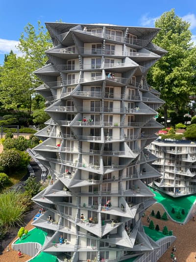 The image depicts a detailed Lego model of a modern, multi-story apartment building with a unique, angular design. The building is constructed from gray Lego bricks, featuring many small balconies and windows. Several Lego figures are placed on the balconies and at the base of the building, suggesting activity. The building is surrounded by greenery, including trees and landscaped areas, also made of Lego. In the background, other Lego structures and trees are visible under a bright blue sky with scattered clouds. This scene is located at LEGOLAND Billund, Denmark, which is a theme park featuring elaborate Lego models of various buildings and landmarks.