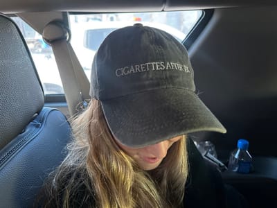 A person wearing a dark-colored baseball cap with the words "CIGARETTES AFTER SEX" embroidered on the front is seated in the back of a car. The person has long, blonde hair and is looking down. A seatbelt is visible over their left shoulder. In the background, a car seat and window are visible with a glimpse of the outside. A water bottle sits on a nearby surface.