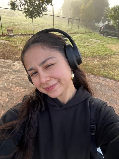A young woman with dark, wavy hair is featured in a selfie, smiling broadly and looking directly at the camera. She is wearing black over-ear headphones and a black hooded sweatshirt. A unique earring, shaped like a miniature ice cream cone, dangles from her ear. Over her left shoulder, a black bag strap with a black buckle is visible, and on it, a small white logo resembling a stylized bird or abstract shape can be seen. She is also wearing a baseball cap under her hoodie, with a white sports team logo visible on its brim.

The setting is outdoors, possibly a park or recreational area in Bassendean, Australia. The woman is standing on a paved walkway made of light brown interlocking bricks. Beyond the path, there is a grassy area with a chain-link fence running through it. In the background, trees are visible, and a dark-colored SUV is parked in the distance. The atmosphere is hazy or foggy, creating a soft, diffused light, suggesting either early morning or late afternoon. The overall mood conveyed by the woman's smile is cheerful and relaxed. There is no clear, readable text in the image other than the aforementioned logos which are not clearly legible as text.