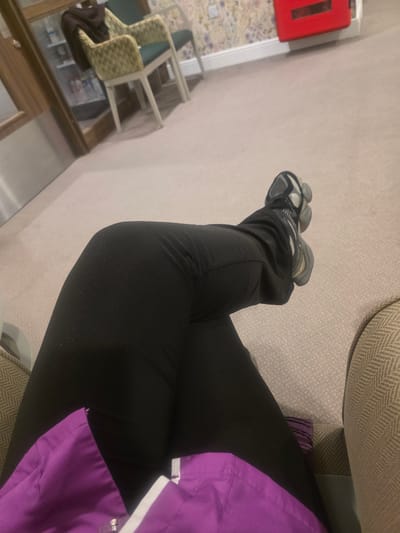 The image captures a first-person view of a person's lower body seated in an indoor setting. The individual is wearing black trousers and a vibrant purple top or jacket, paired with grey and black athletic shoes. Their legs are crossed at the ankles.

The scene appears to be a public or semi-public waiting area or common room. The floor is covered with a light-colored, likely beige, carpet. The person is seated on an upholstered piece of furniture, partially visible on the right, featuring a beige fabric with a distinct herringbone pattern.

In the background, to the left, two additional chairs are visible, both with light-colored frames and patterned upholstery, in shades of green and yellow. Below these chairs, there's a dark, reflective surface, possibly a table or a mirrored panel. Mounted on a light-colored wall further to the left is a prominent red rectangular object, resembling a fire alarm pull station or an emergency box.

The only activity depicted is the person sitting. The time of day is not discernible due to indoor lighting. No discernible text is visible on any objects in the image. The environment suggests an institutional or commercial building, consistent with an indoor public space in Harrogate, United Kingdom.