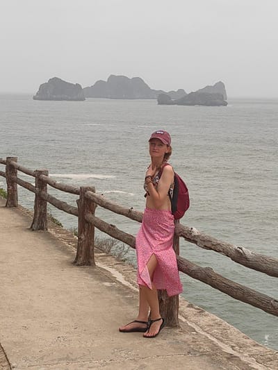 A photograph shows a woman standing next to a wooden railing, with the ocean and islands in the background. The woman is wearing a red baseball cap, a floral-patterned bikini top, a red backpack, a pink floral sarong-style skirt, and black sandals. She is leaning against the railing, and appears to be posing for the photo. The islands are in the distance, shrouded in mist. The railing is made of unfinished wood and appears to be a safety barrier along a concrete path. The scene is likely taken on Cat Ba Island, part of the city of Hải Phòng, Vietnam.