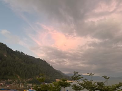 The image depicts a scenic view of a mountainous landscape and a body of water, likely a lake, in Mendale, Indonesia. The sky is a prominent feature, with dramatic clouds that are tinged with pink and orange hues, suggesting either a sunrise or sunset. A large, tree-covered mountain slopes down from the left side of the frame towards the water. At the base of the mountain, there are some buildings and structures, including what appears to be a dock or pier with several boats moored. In the foreground, several trees with sparse foliage are visible, framing the view of the water and the distant mountains across it. The overall atmosphere is serene and natural, with a hint of human presence in the settlement by the water.
