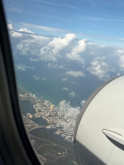 The image shows a view from an airplane window. The wing of the plane is visible in the upper right. Below the wing, there's a partial view of what appears to be the city of North Miami, Florida, USA. The coastline is visible, with high-rise buildings lining the shore. There are also inland waterways and forested areas. Above the coastline, there are scattered clouds in the sky.
