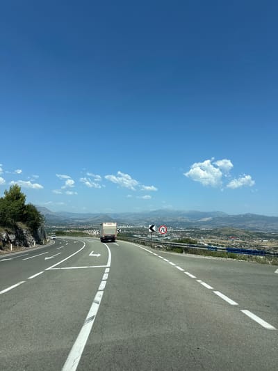The image shows a scenic view of a highway in Gornji Kokoti, Montenegro, with a truck in the foreground and a town in the background. The sky is clear blue with a few scattered clouds. The highway curves to the left, with white lane markings and directional arrows painted on the asphalt. A speed limit sign indicating 70 km/h is visible on the right side of the road. In the distance, a town is nestled among rolling hills and mountains. The overall scene depicts a bright, sunny day in a picturesque Mediterranean landscape.
