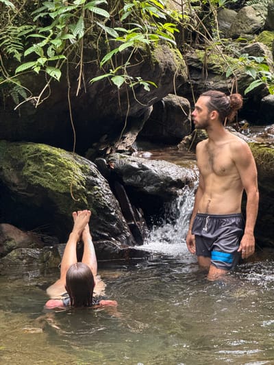 A man with long hair pulled into a bun, wearing dark shorts with blue accents, is waist-deep in a stream or river. He leans forward, partially submerged in the water. Opposite him, a person with long hair is face-down in the water, hands extended. The setting appears to be a natural area, likely Suối Giàng, Vietnam, with moss-covered rocks and lush vegetation surrounding the water. The water is clear, with rocks visible beneath the surface.