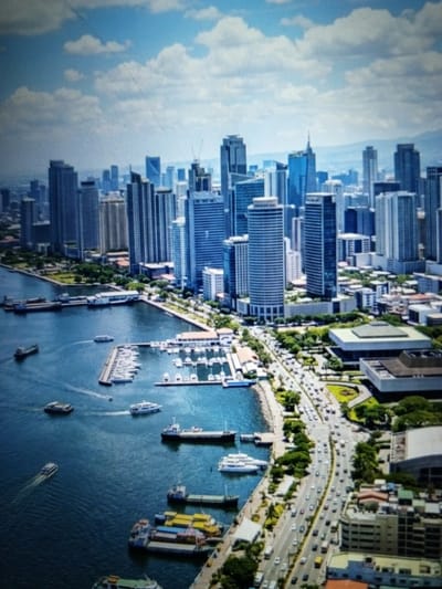 This aerial photograph depicts a bustling cityscape with a prominent waterfront. Tall, modern skyscrapers dominate the background, their glass facades reflecting the bright blue sky and scattered white clouds. In the foreground, a large body of blue water is filled with various vessels. Several boats are docked at a marina, while others navigate the open water, leaving wakes behind them. A busy road runs parallel to the waterfront, lined with trees and more buildings. The scene suggests a vibrant urban environment with significant maritime and commercial activity. This image is not of Golbembal, Nigeria.