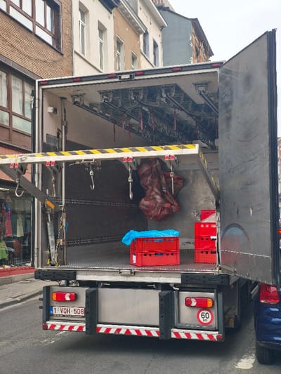 A large, white delivery truck is parked on a street in Saint-Josse-ten-Noode, Belgium. The back of the truck is open and a large piece of meat is hanging from the ceiling. The truck is facing forward, and its license plate number is 1 VOH-508. The truck is parked next to a brick building with windows. There are two red plastic crates inside the truck. The street is paved with gray asphalt. The truck has red taillights and a 60 km/h speed limit sign. There is a small section of a blue car visible in the lower right corner of the image.