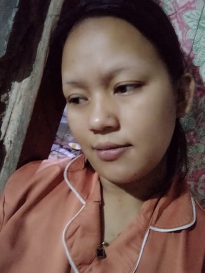 A young woman with dark hair pulled back poses in a close-up shot. She has smooth, tan skin and is wearing a salmon-colored pajama top with white piping. A gold necklace with a dark pendant hangs around her neck. She looks to the left with a slight, almost introspective expression. Behind her, a wall is partly visible, with a combination of a wooden structure on the left and patterned fabric on the right.