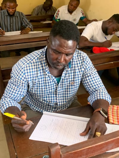 The image captures five adult Black males seated at individual dark wooden desks within an indoor room, identified as being in Abobo, Nigeria. The foreground subject, a male with short dark hair and a beard, wears a blue and white checkered long-sleeved shirt over a light blue garment. He is leaning forward, looking down, and actively writing on a white lined notepad with a yellow pen held in his right hand. A brown-strapped smartwatch is present on his left wrist, and a gold-colored signet ring is on his right ring finger. A multi-colored plaid cloth is partially visible on his desk to the right. In the background, four other adult Black males are similarly seated at wooden desks, also focused on writing on papers. Their attire includes a striped long-sleeved shirt, a grey shirt, a white polo shirt with green and white accents, and a white t-shirt. A red cap rests on one of the desks in the background. The room's walls are light-colored, suggesting a classroom or training setting.