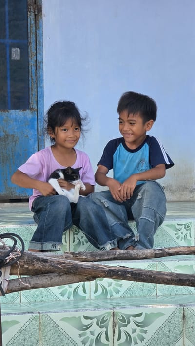 Two children, a boy and a girl, are outside in what appears to be Ba Ngoi Ward, Vietnam. The boy, wearing a blue and white t-shirt and jeans, kneels next to the girl. He is smiling and reaching towards the girl's hands. The girl, wearing a pink t-shirt and jeans, is holding a black and white cat in her arms. She is also smiling. They are on tiled floor with floral patterns next to a blue metal door and some rough wooden poles.