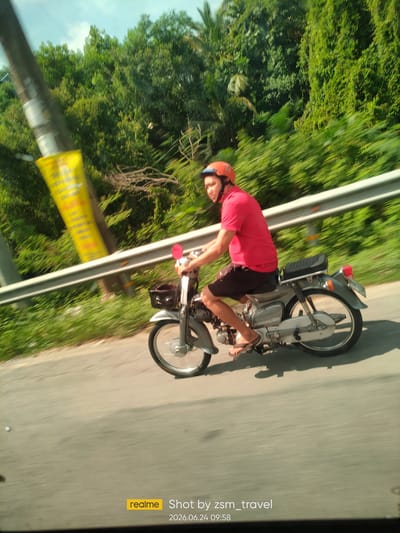 A person is riding a motorcycle on a road in Ben Cau, Vietnam. The person is wearing a red shirt, black shorts, and an orange helmet. They are riding on the right side of the road, and there is a metal guardrail to their left. The background is filled with lush green trees and vegetation. The sky is cloudy. The image is a bit blurry due to motion. A "realme" watermark and the date and time "2026.06.24 09:58" are visible in the bottom right corner.