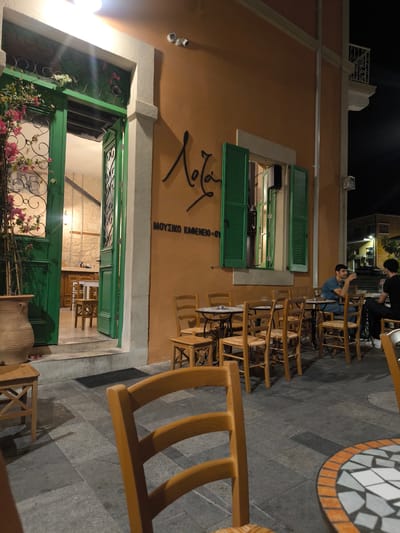 This image depicts the exterior of a restaurant named "Lola" in Paphos Municipality, Cyprus, during nighttime. The restaurant has a warm, orange-colored facade with a green-painted doorway that is partially open, revealing the interior. The doorway is adorned with intricate metalwork. Above the doorway, there is a white decorative molding. To the right of the entrance, a large window with green shutters is visible, with a black speaker mounted above it. A sign with the name "Lola" is written in stylized script on the building's wall, accompanied by Greek text reading "ΜΟΥΣΙΚΟ ΚΑΦΕΝΕΙΟ-ΟΥ" (Music Cafe).
The outdoor seating area is furnished with numerous wooden chairs and small, round tables. Two men are seated at one of the tables, engaged in conversation. The man on the left is wearing a blue t-shirt and appears to be holding a drink. The man on the right is wearing a black t-shirt. The ground is paved with large, grey tiles. A large terracotta pot containing a plant with pink flowers stands to the left of the entrance. Another wooden chair is prominently featured in the foreground, partially obscuring the view of the tables and the street. The overall atmosphere suggests a casual dining or cafe setting at night.
