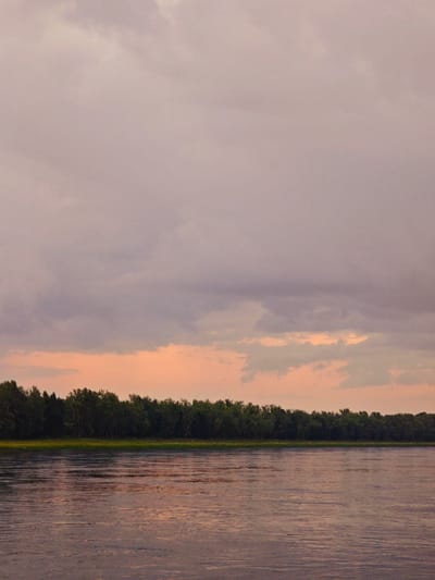 An expansive, calm river stretches across the lower half of the frame, reflecting the muted colors of the sky. The water surface is smooth with subtle ripples. Along the far bank, a dense line of dark green trees forms a natural boundary, their foliage creating a textured silhouette against the lighter horizon. Above the treeline, a band of pale orange light suggests the setting or rising sun, diffused by a layer of soft, hazy clouds that dominate the upper portion of the image. The overall atmosphere is serene and tranquil. The location is a riverbank in Kazachinskoe, Russia.