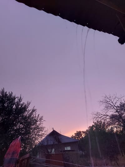 The image captures a twilight sky in Dachi Su, Kyrgyzstan. The sky transitions from a deep purple near the top left to a soft pink and orange glow on the right, indicating sunset or sunrise. In the foreground, a dark, textured surface, possibly a roof or overhang, occupies the left side of the frame. On the right, silhouettes of trees and a building with a corrugated metal roof are visible against the colorful sky. The building has a window that appears to be illuminated from within. Faint, wispy clouds or contrails are visible horizontally across the sky. The overall mood is serene and tranquil, with the soft light of dusk casting a peaceful atmosphere over the scene. The image is oriented vertically, with the sky filling the majority of the frame.