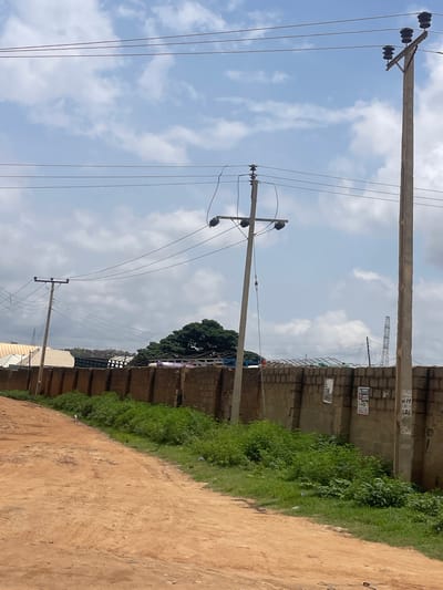 The image shows a dirt road in front of a low brick wall in Unguwar Saya, Nigeria. Electrical poles and wires run across the sky above the wall. A large green tree is visible behind the wall, and several buildings with white roofs are in the distance. The sky is cloudy.