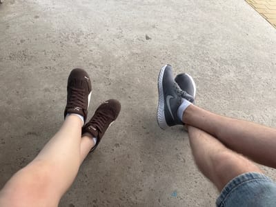 Two pairs of legs are visible, resting on a grey concrete surface. The left legs are wearing brown Puma sneakers with white soles and white socks. The right legs are wearing grey Nike sneakers with white soles and white socks. One person's legs are crossed at the ankles. The concrete is textured and appears to be outdoors, with a hint of a yellow brick-like surface visible in the upper right corner. The lighting suggests it is daytime. No specific location in Shulhynka, Ukraine can be identified from the image alone.