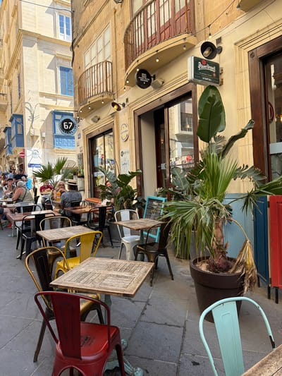 This is a ground-level, eye-level, medium shot of a bustling outdoor cafe in Valletta, Malta. The scene is oriented vertically, suggesting it might have been taken with the camera rotated. The sun casts strong shadows, indicating it's likely midday. The cafe is set up on a cobblestone street lined with traditional Maltese architecture, characterized by pale yellow stone buildings and ornate balconies.
Several tables and chairs are arranged on the sidewalk, with some patrons seated and others implied to be nearby. The people are dressed casually, some appear to be tourists. A few tables have glasses of drinks and patrons are engaged in conversation, suggesting a relaxed social atmosphere.
Prominent objects include outdoor cafe furniture, a large potted plant with lush green foliage, and signage. Visible text includes "Pilsner Urquell THE ORIGINAL PILSNER," "67 KAPITALI," "CITY CENTRE," and "MAGIC ROCK BREWING." There are also decorative light strings adding to the ambiance. The overall impression is one of a lively European street scene during daylight hours.