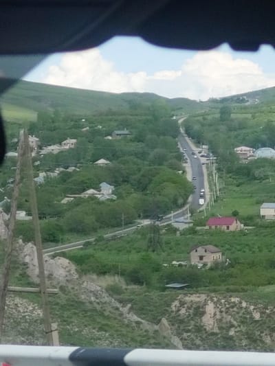 The image displays an elevated, slightly angled view of a village or settlement nestled within a verdant, hilly landscape, characteristic of Zangakatun, Armenia. A winding asphalt road is a prominent feature, traversing the valley or hillside, with several vehicles—including cars and a white van—visible on it, indicating ongoing traffic.
Numerous houses of varying sizes and architectural styles are scattered across the lush green terrain; some boast distinctive red or dark roofs, while others appear lighter. In the upper-left section, a large, light-colored domed building can be seen. The surrounding environment is dominated by dense green foliage, trees, and grass covering the hillsides, interspersed with some exposed rocky or dirt patches.
A bright blue sky with fluffy white clouds is visible on the left, suggesting clear, daytime weather conditions. In the immediate foreground, dark, out-of-focus elements on the left side suggest the viewpoint is from inside a vehicle or through a window, possibly a silhouette. On the right, a vertical white and grey structure, potentially a railing or post, and some rustic wooden fence-like elements are partially visible, framing the scene.
The image captures a peaceful rural or semi-rural setting, depicting daily life and infrastructure integrated with the natural topography. There is no visible text in the media file.