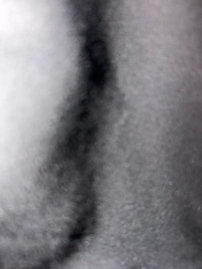 A blurry, abstract image presents a field of gray tones, punctuated by a dark, meandering line. The texture appears to be somewhat granular, creating a diffused, out-of-focus effect. The dark line weaves irregularly across the frame, contrasting with the lighter gray areas. The overall impression is atmospheric and indistinct, lacking sharp details or clear forms.