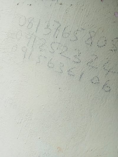 The image depicts a close-up view of a textured, light-colored wall, possibly painted concrete or plaster. Faint, blue-ink markings, appearing to be numbers or letters, are scrawled across the surface. The markings are inconsistent in clarity and form, suggesting they may have been written with a pen or crayon. Some are clearer than others, with distinct shapes of what appear to be digits like "0", "1", "5", "7", "8", "2", "3", "6", and "9". There are also partial markings and smudges. The lighting is even, with no strong shadows, indicating ambient or diffused light. The surface of the wall is rough and uneven, with small pockmarks and a few larger, darker blemishes. There are no visible people, animals, or other distinguishing objects in the frame. The context of Gindin Dutse, Nigeria, is not visually represented in the image itself, as there are no identifying landmarks or elements that would place the scene geographically. The image focuses solely on the wall and its markings.