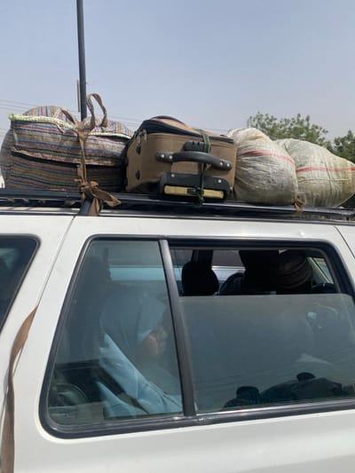 This image depicts the side view of a white vehicle, likely an SUV, with a loaded roof rack, situated in Gobirawa, Nigeria, under clear, bright daylight.

**Objects:**
*   **Vehicle:** A white four-door SUV is positioned with its right side facing the viewer. The rear side windows are tinted but allow for partial visibility of occupants.
*   **Roof Rack Contents:**
    *   A large, multi-colored striped fabric bag, secured with a brown fabric strap, is positioned on the front left of the roof rack.
    *   A brown wheeled suitcase, with its handle retracted, is secured with a green strap or bungee cord next to the striped bag.
    *   Two white woven sacks, each featuring red stripes, are placed behind the suitcase, towards the rear of the roof rack. All items are firmly affixed to a black roof rack structure.
*   **Environment:** The sky is a light blue, almost white, suggesting an intense sun. In the upper left, a dark, thin vertical pole extends upwards. In the distant background on the right, sparse green foliage from trees is visible against the light sky.

**People:**
*   **Rear Left Passenger:** A person is visible through the tinted rear left passenger window. They appear to be wearing a light-colored garment, possibly a head covering, and are seated, with their face partially obscured.
*   **Rear Right Passenger:** Another individual is visible through the rear right passenger window, wearing a dark cap or traditional headwear and dark clothing. Their face is also largely indistinct.
*   Other occupants may be present but are not clearly discernible through the vehicle's windows.

**Actions:** The vehicle appears to be stationary or in transit, laden with luggage on its roof rack, indicating travel or preparation for travel. The people are seated within the vehicle.