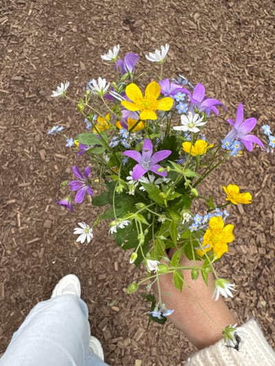 A person wearing light blue pants and a cream-colored sweater holds a bouquet of wildflowers. The bouquet consists of various colored flowers, including yellow buttercups, purple bell-shaped flowers, small white flowers, and tiny blue forget-me-nots. The flowers are arranged in a slightly messy, naturalistic style. The person's hand, with a visible watch on the wrist, is holding the stems of the bouquet. Their feet, clad in white sneakers, are visible in the lower left corner of the image. The background is a surface covered with wood chips, suggesting an outdoor setting, possibly a park or a garden path. The lighting suggests it is daytime. No specific landmarks of Bakhtiyary, Russia, are visible in the image.