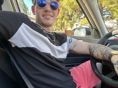 A male individual is seated in the driver's seat of a vehicle, looking towards the camera and smiling. He wears a black and white horizontal striped t-shirt, pink shorts, and round, reflective blue-tinted sunglasses. A silver chain necklace with a small blue pendant is visible. Both arms display extensive tattoos; the left forearm features multiple clock faces, while the right forearm shows a skeletal figure. A gold-toned wristwatch with a brown strap is on his left wrist. His right hand rests on the black steering wheel. The driver's side seatbelt is visible but not clearly buckled across his chest. Through the vehicle's windows, a residential street in Sparks, Nevada, United States of America, is visible under clear daylight conditions. Trees with green and yellow foliage, a light-colored house, and a wooden fence are reflected in the side mirror and visible through the windshield.